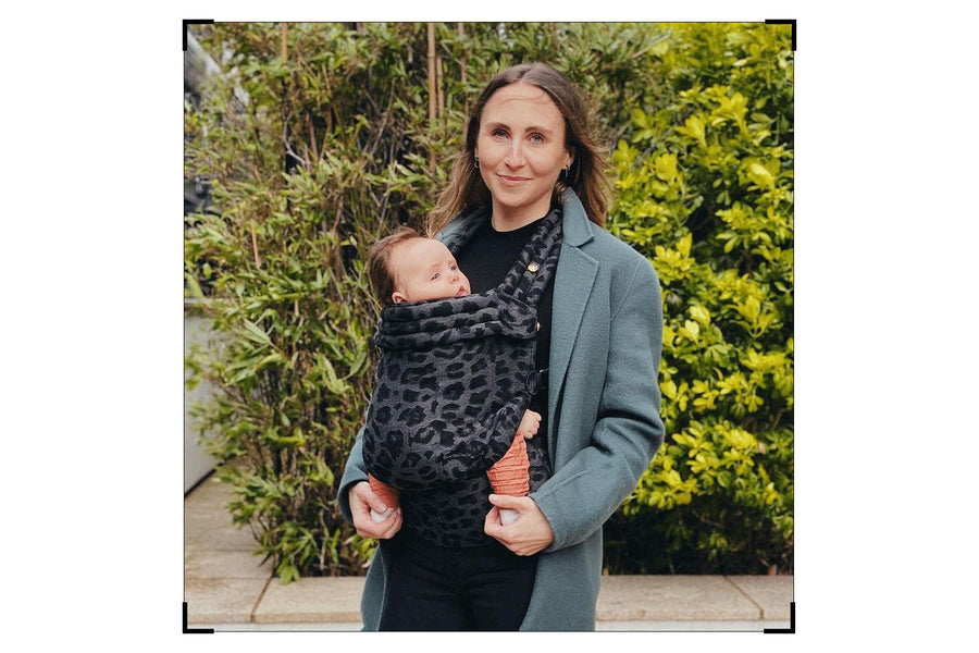 Rachel holds her baby in a leopard print carrier. The baby, dressed in an orange Versatile set by Petit Pli, is nestled close against their chest. The image captures a tender parenting moment.