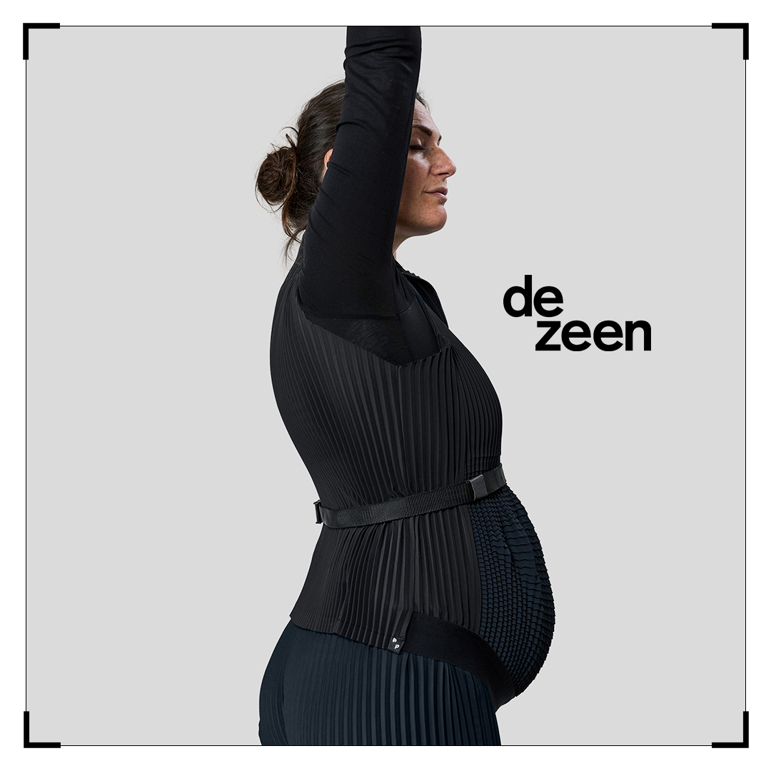 Pregnant model in sculptural black Petit Pli outfit, arms lifted in poise, framed by Dezeen logo in a minimalist, editorial composition