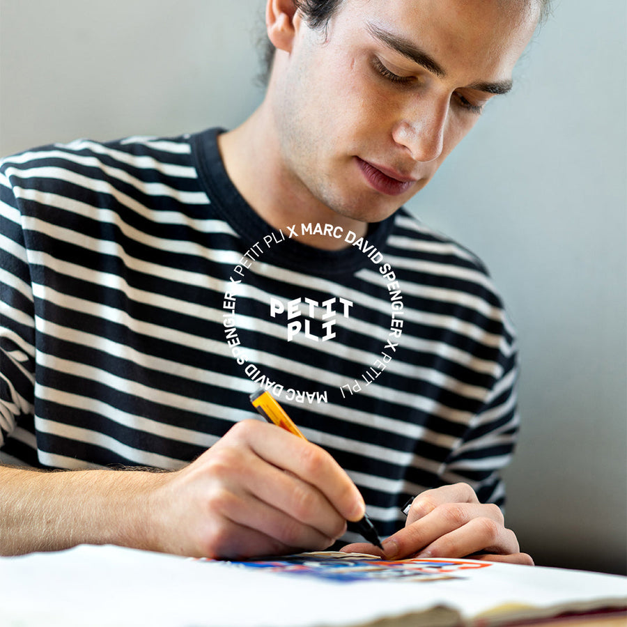 Marc sat in a striped shirt sketches on paper, framed by circular Petit Pli x Marc David Spengler text, highlighting their Print 08 creative collaboration.