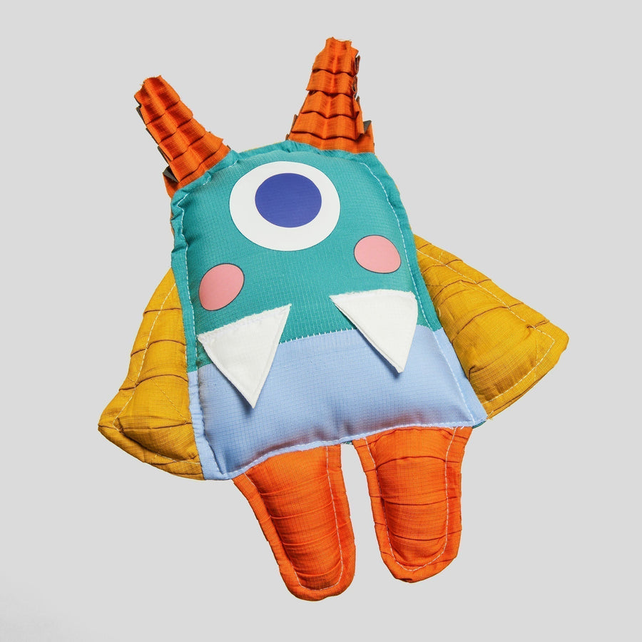 Ivy the Petit Pli mascot with one large eye, orange horns, yellow wings and orange legs, handcrafted from recycled offcuts to inspire MiniHuman adventures.