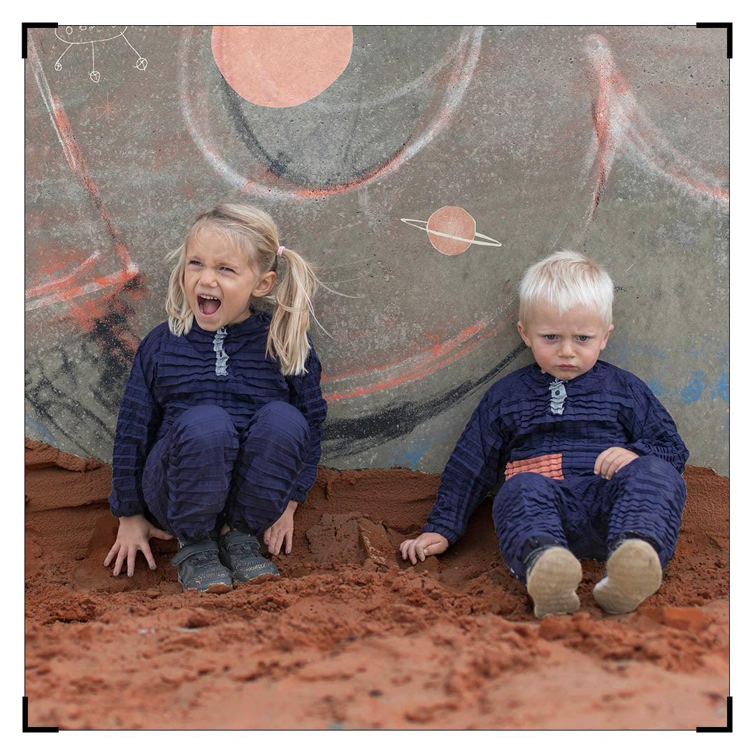Two LittleHumans in dark blue Petit Pli outfits sit on sand before a space‑themed mural, capturing the spirit of Clothes That Grow and Mars Test play.