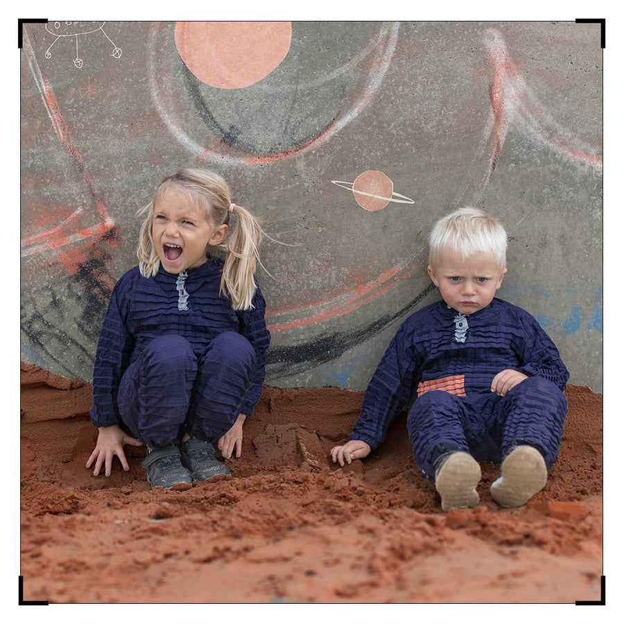 Two LittleHumans in dark blue Petit Pli outfits sit on sand before a space‑themed mural, capturing the spirit of Clothes That Grow and Mars Test play.