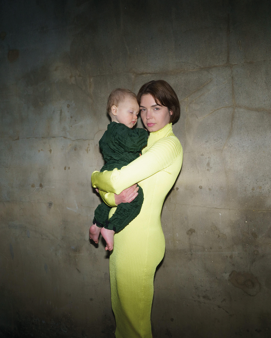 Nagyedi in a lime green dress stands holding her LittleHuman Mika who is wearing one of Petit Pli's first ever Green Ore Versatile Suits
