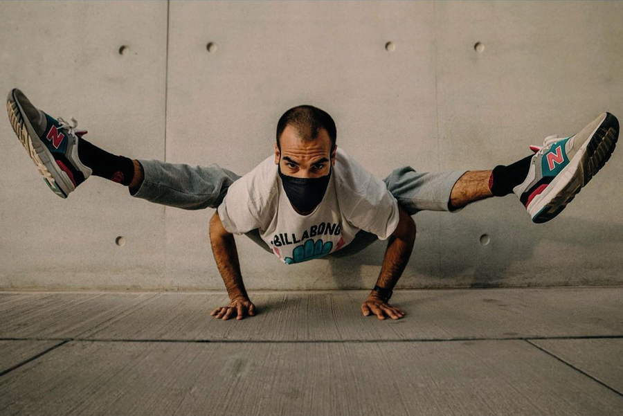 Yoga guru Panagiotis Kontis balances in a horizontal handstand, wearing Eclipse Black mask, white tee, blue trousers, against a plain concrete wall