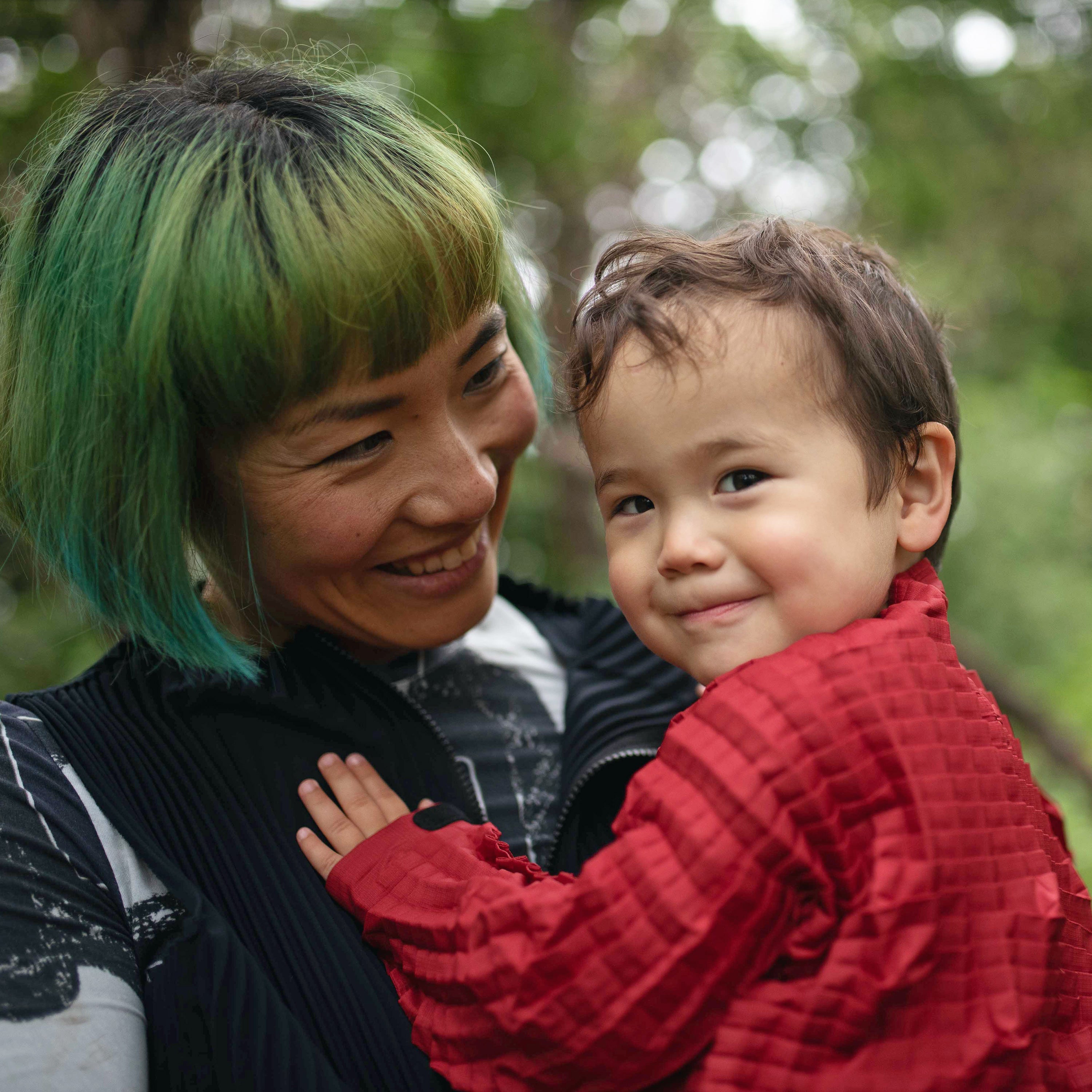 Masumi holds LittleHuman Towa outdoors, both smiling gently. Towa wears a red Petit Pli jacket as they share a quiet moment in nature.