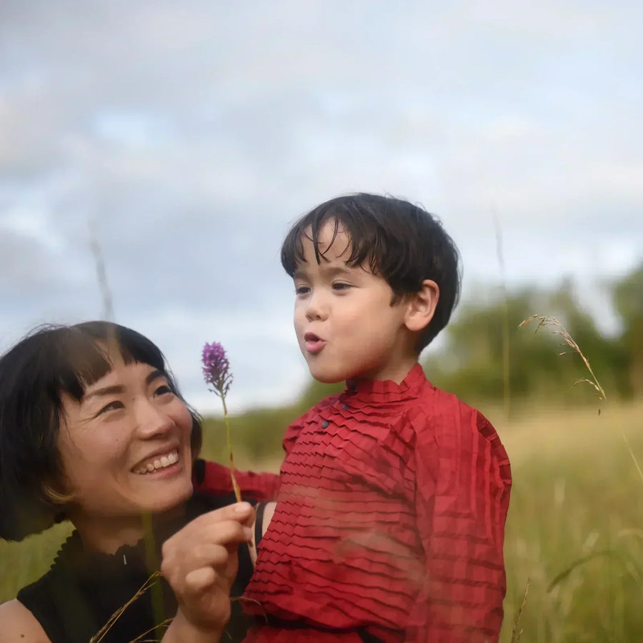 A mother and child share a moment in a grassy field, holding a flower, reflecting Petit Pli’s focus on curiosity, bonding, and emotionally durable design.