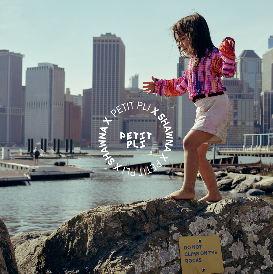 A child balances barefoot on a rock by the water in a colourful Petit Pli x Shawna X outfit, set against a sunny city skyline and playful backdrop of New York.