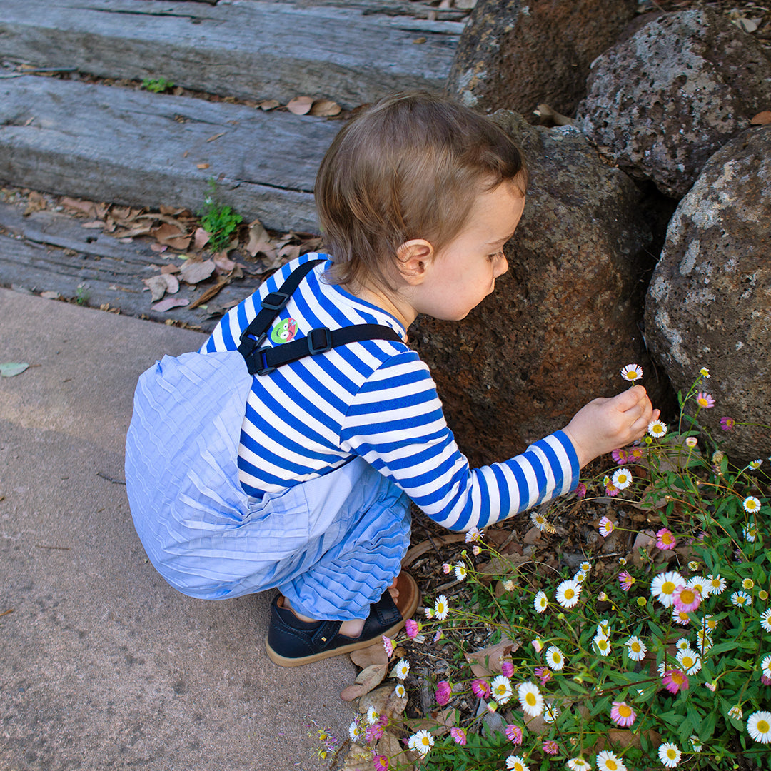 A child in striped shirt and Versatile Dungarees crouches to touch flowers outdoors, reflecting Petit Pli’s Clothes that Grow for everyday exploration.