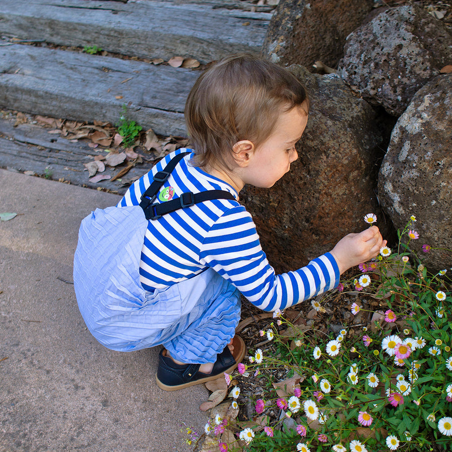 A child in striped shirt and Versatile Dungarees crouches to touch flowers outdoors, reflecting Petit Pli’s Clothes that Grow for everyday exploration.