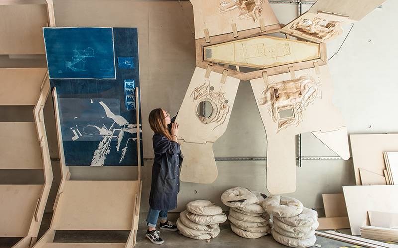 Person viewing a large abstract wooden installation with blueprints in a London art studio, reflecting creativity and innovation in student design shows.