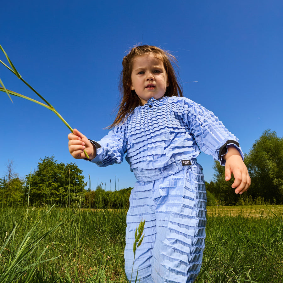 A child stands in a grassy field wearing Petit Pli’s expandable outfit, holding a plant stem capturing movement, growth, and outdoor play in frame.